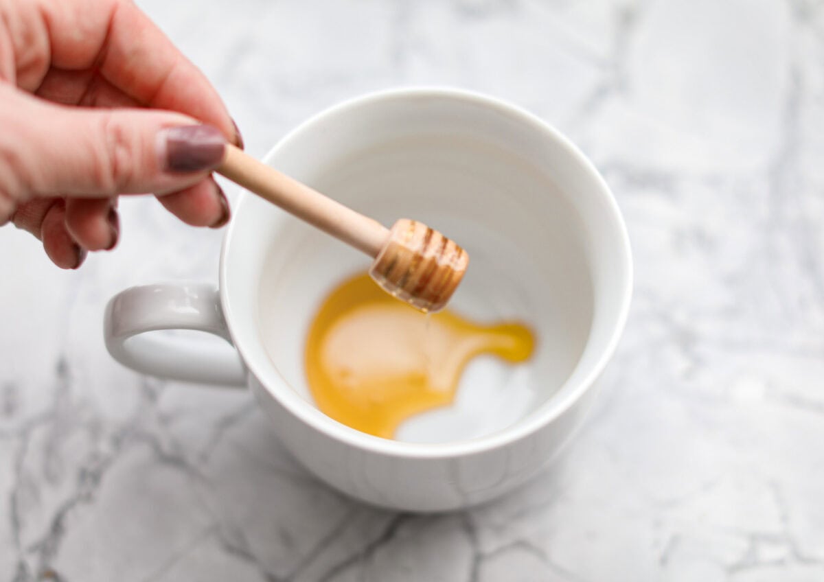Whenever I am sick, the Starbucks Medicine Ball is my go-to, and this homemade version is even better! Perfect for cold and flu season, this warm drink made of lemonade, honey, and tea is soothing and delicious! Overhead shot of someone putting honey in the bottom of a mug.